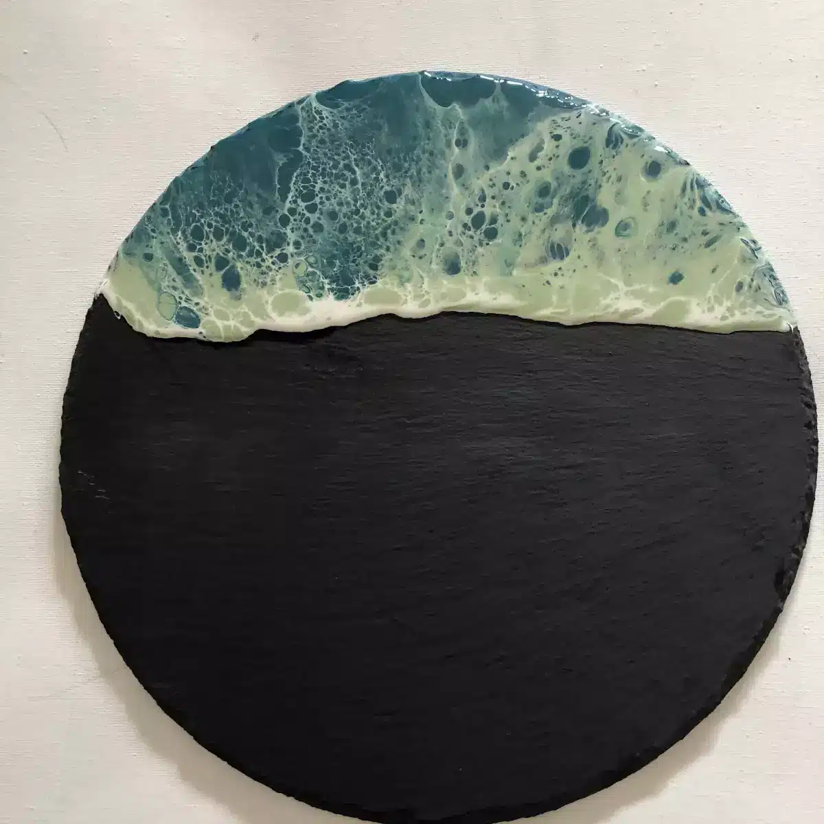 Art en résine style vague océanique turquoise et blanche sur un plateau circulaire en ardoise noire texturée.