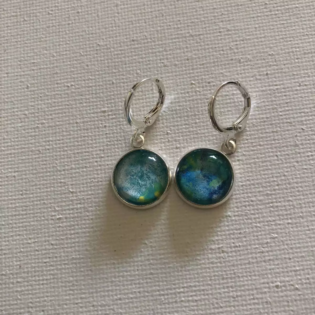 Boucles d'oreilles pendantes rondes avec motifs abstraits bleu et vert, monture argentée sur fond blanc texturé.