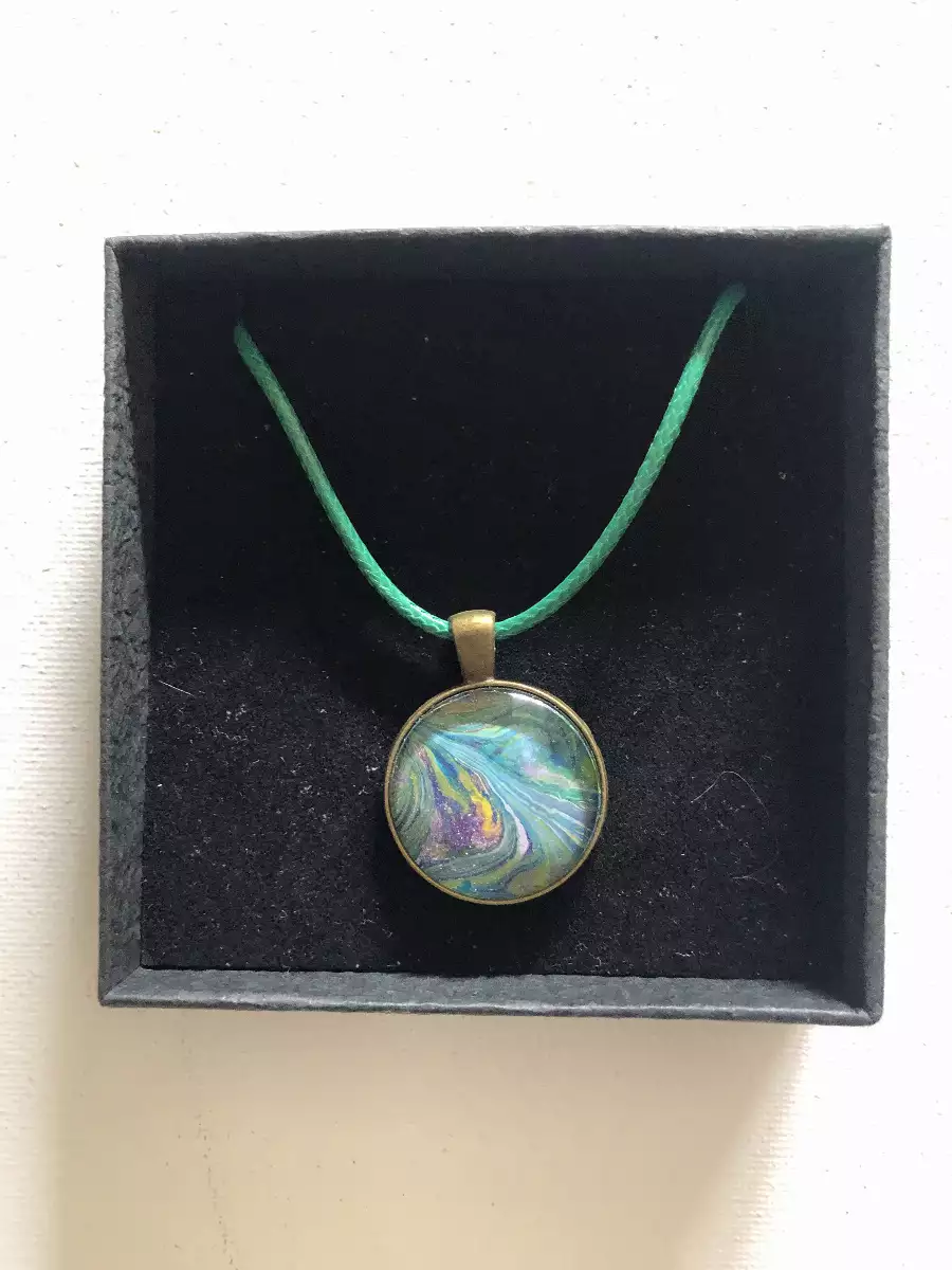 Pendentif rond en peinture fluide bleu et vert sur cordon assorti dans son écrin noir.