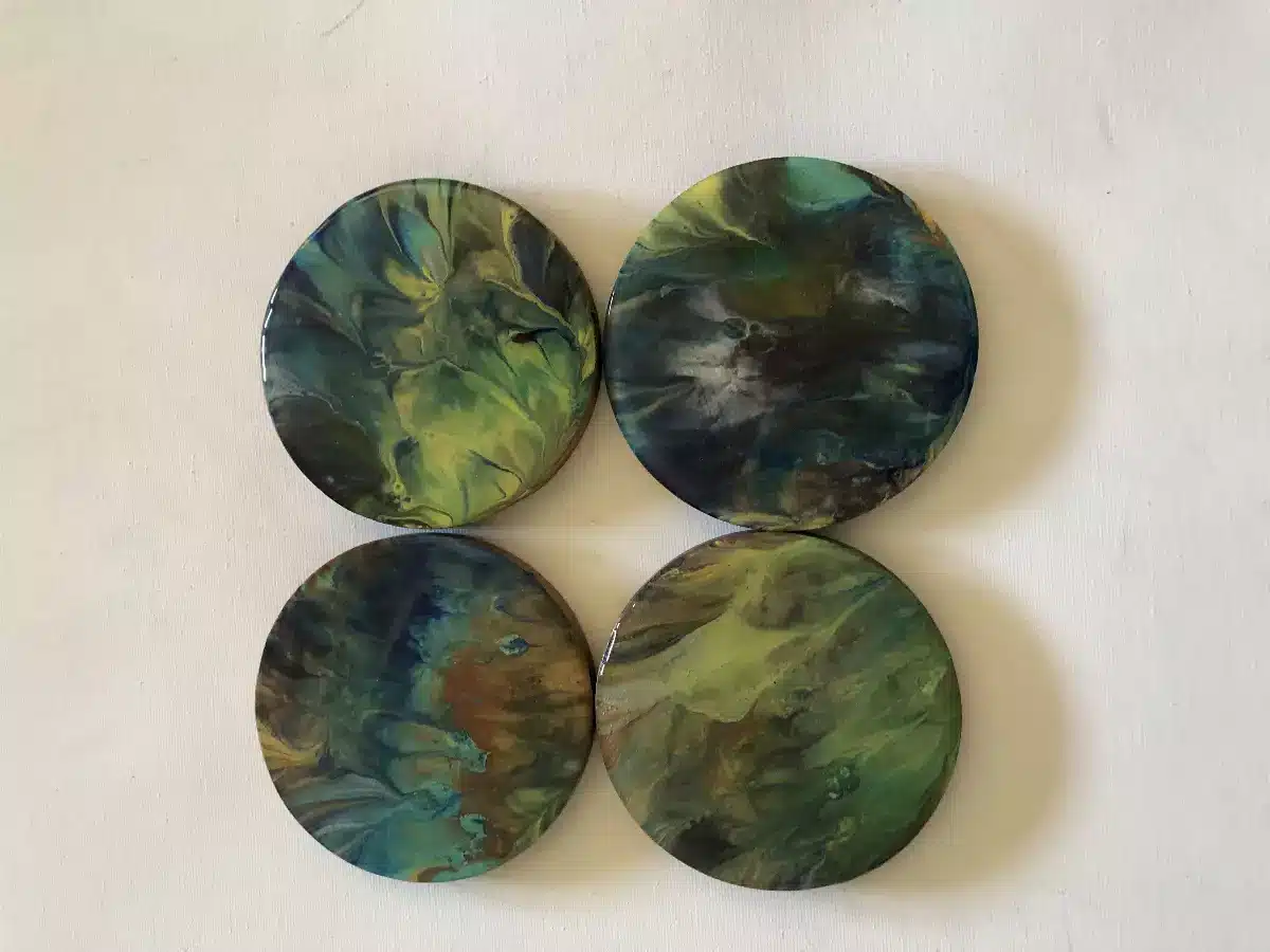 Quatre dessous de verre ronds en art fluide avec motifs abstraits verts, bleus et jaunes sur fond blanc.