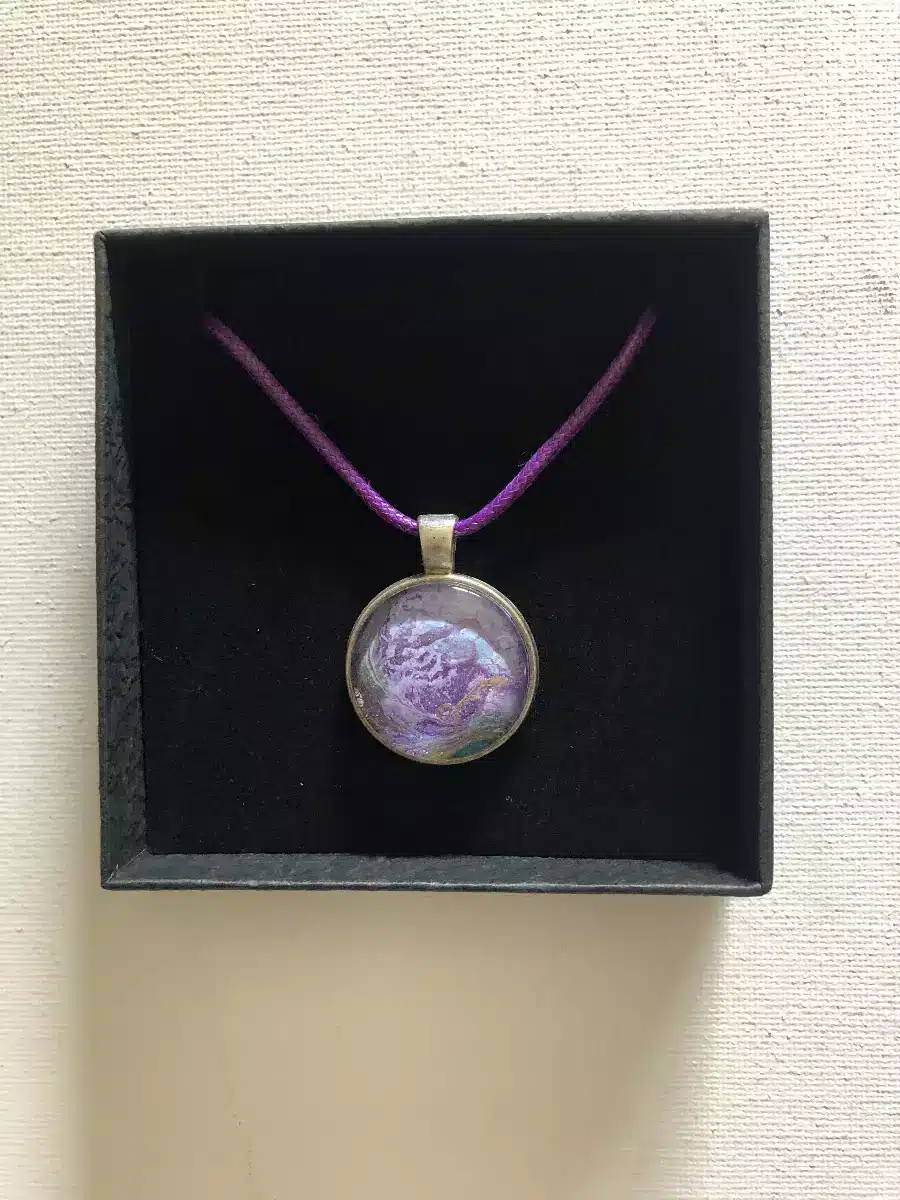 Pendentif rond aux nuances violettes et dorées sur un cordon violet, présenté dans son coffret cadeau noir.
