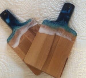 Deux planches à découper en bois ornées d'une résine époxy bleue et blanche évoquant des vagues sur le sable.