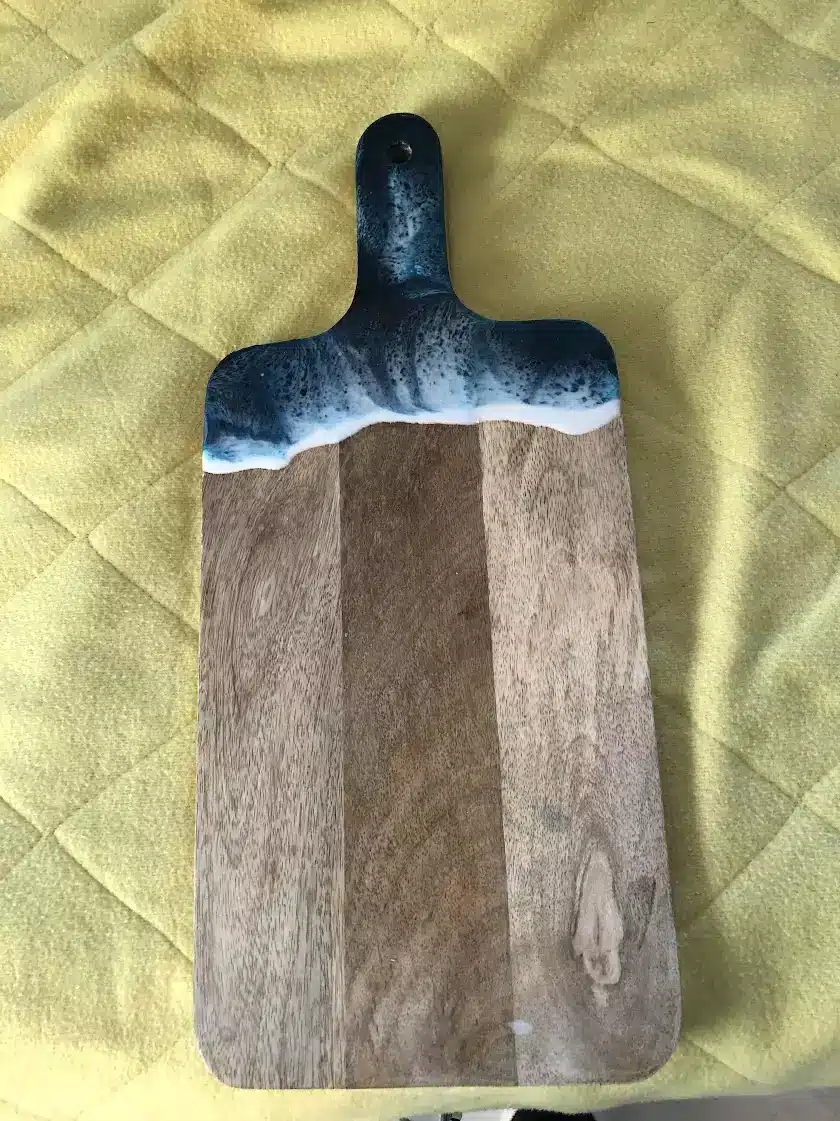 Planche à découper en bois avec décor de vagues en résine époxy bleue et blanche, posée sur un tissu jaune matelassé.